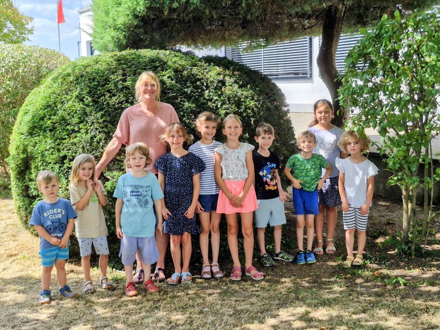 Gruppenbild der Kichererbsen: zehn Kinder und ihre Chorleiterin stehen im Sommer vor einem runden, dicht geschnittenen Busch